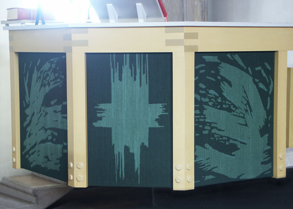 Close-up of a church pulpit decorated with green woven panels showing abstract patterns framed by light wood supports.