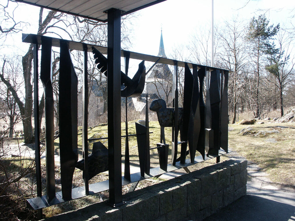 Black metal sculpture with abstract vertical forms and a stylized animal figure, mounted outdoors on a low stone wall with trees in the background.