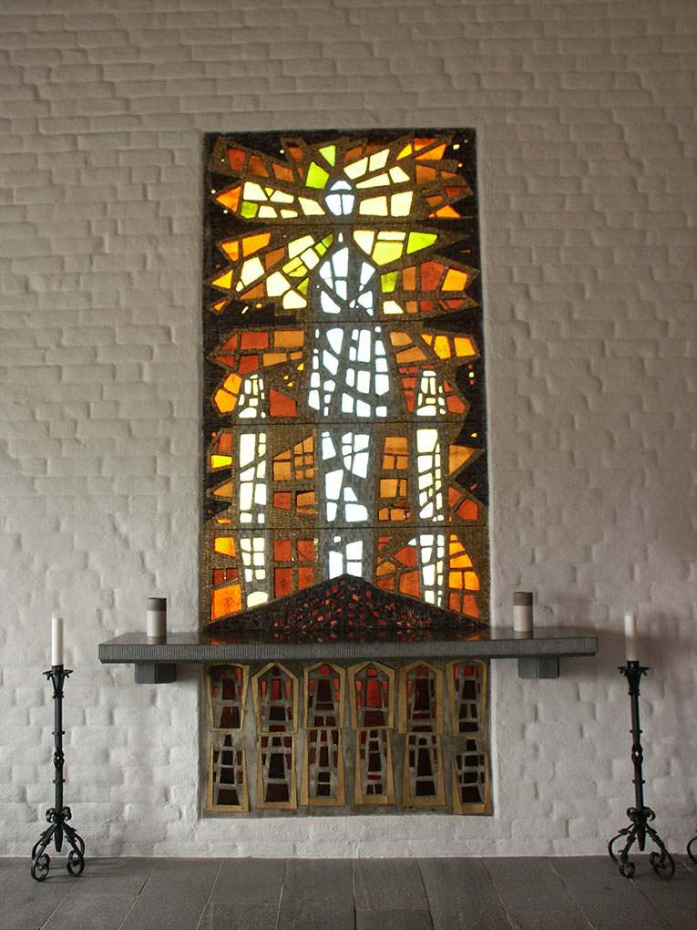 A stained glass window in a brick chapel showing abstract tall radiant figure above the altar with candles and metal stands.