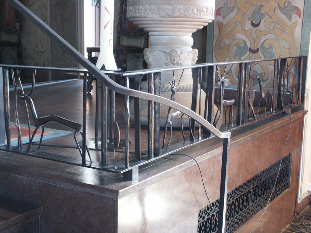 Wrought-iron church railing with stylized animal figures near a carved stone baptismal font and patterned wall paintings in soft light.