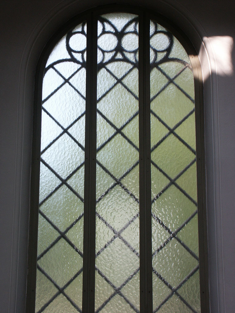 Tall arched window with textured glass panes arranged in a diamond pattern and decorative circular metalwork near the top.
