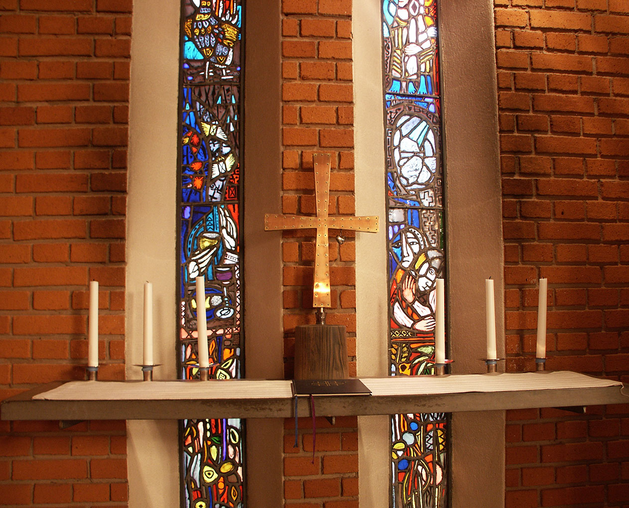 Brick-walled church altar with a metal cross, white candles, and three narrow stained-glass windows depicting colourful religious scenes.
