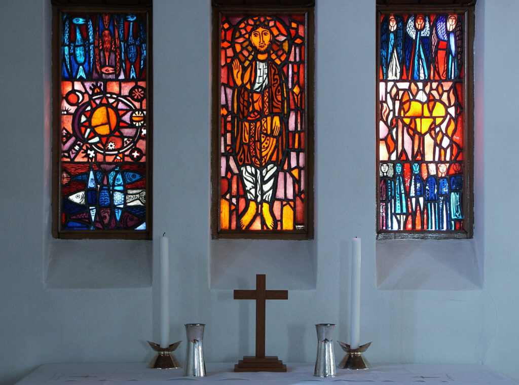 Three vibrant stained-glass windows depicting symbolic scenes flank a simple altar with a wooden cross and tall white candles.