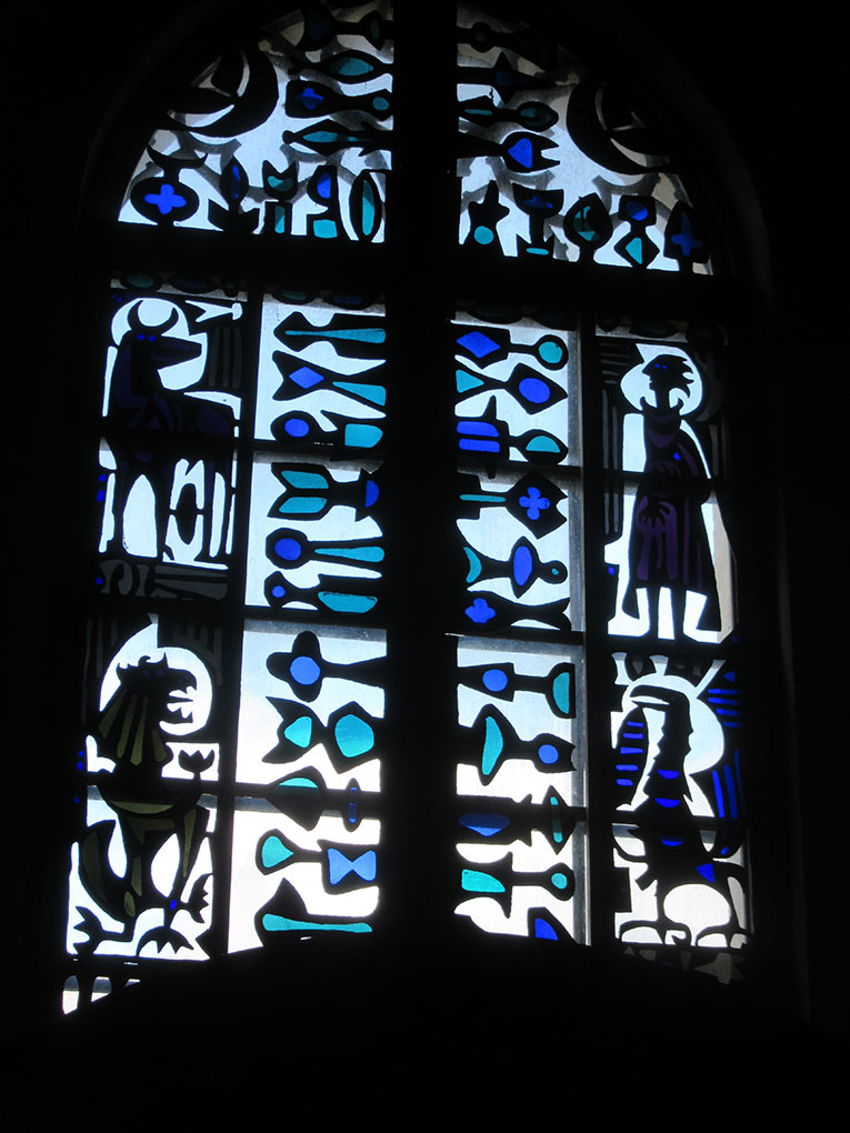 Arched stained-glass window with blue and teal abstract fish motifs, human figures, and symbolic shapes illuminated against a dark interior.