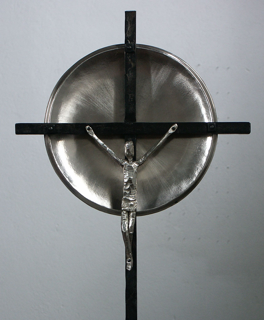 Metal crucifix sculpture with a textured figure on a simple cross set before a round, reflective metal disc against a plain grey background.