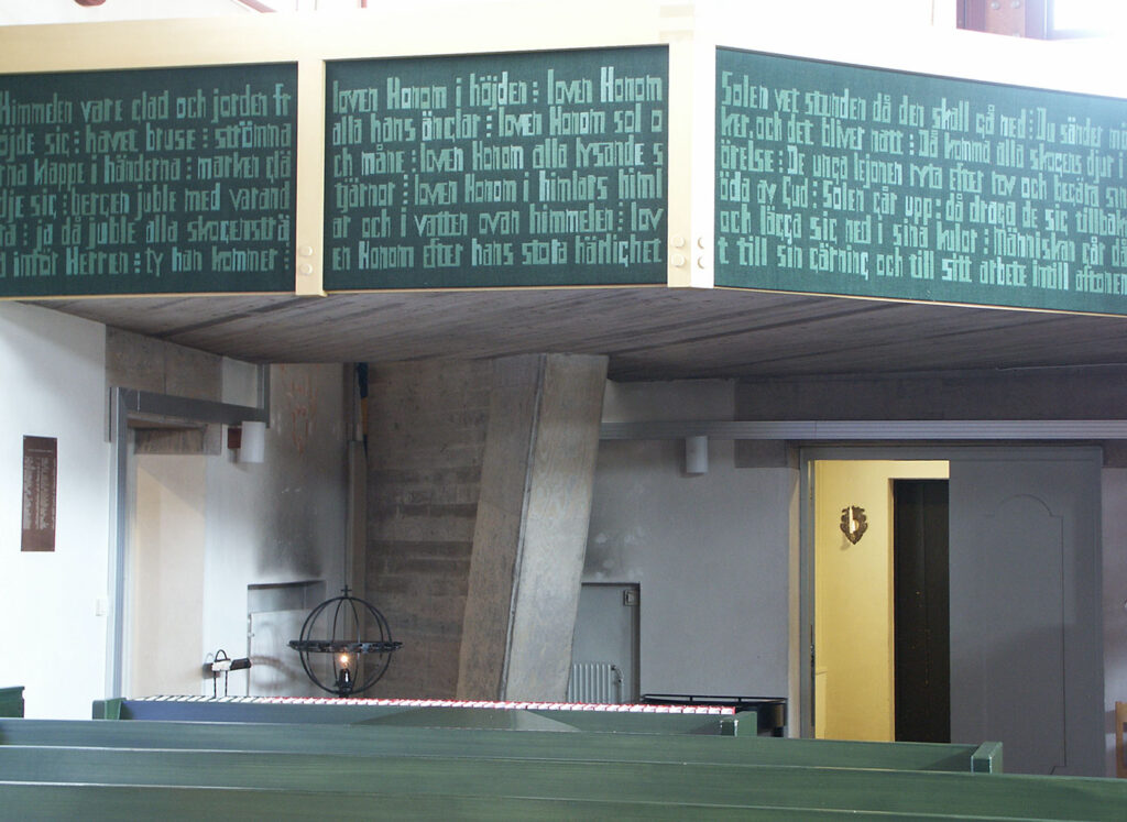 Green scripture panels wrap around the church balcony above green pews, with a candle holder and doorway visible beneath the concrete supports.