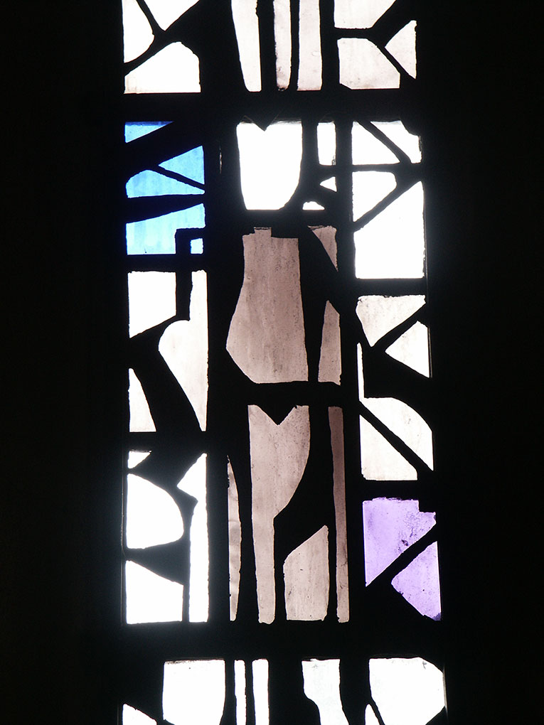 Close-up of abstract stained-glass window with angular black lead lines framing white, blue, and purple glass shapes glowing in sunlight.