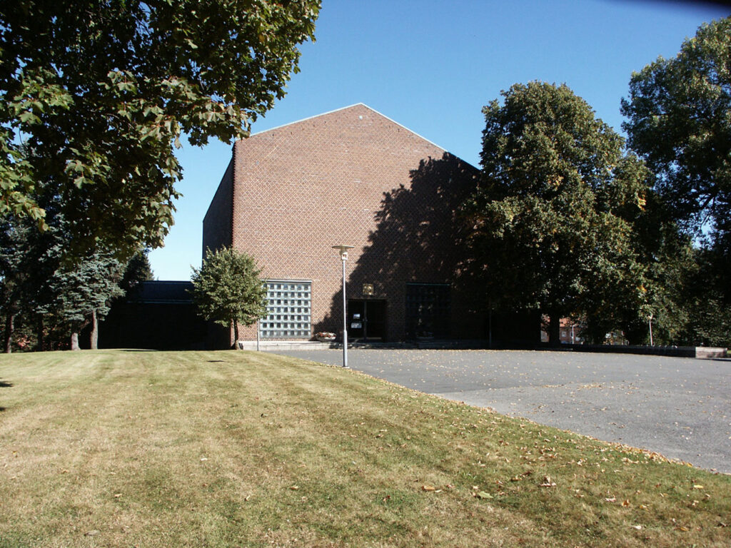 Large brick building with a steep roof, glass-block windows, surrounding trees, and a wide lawn under a clear blue sky.
