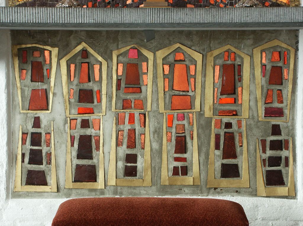 Abstract stained-glass panel with geometric red and orange shapes set in tall, narrow frames above a cushioned surface against a textured wall.