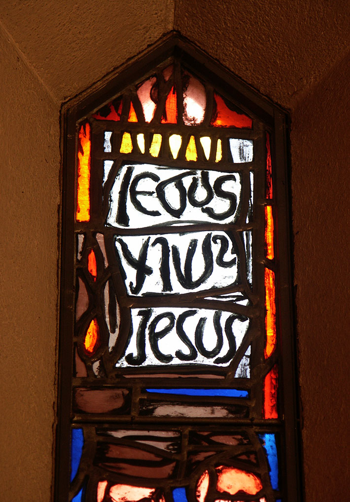 Narrow stained-glass window with warm red, orange, and blue tones, featuring abstract shapes and the repeated handwritten name “Jesus.”