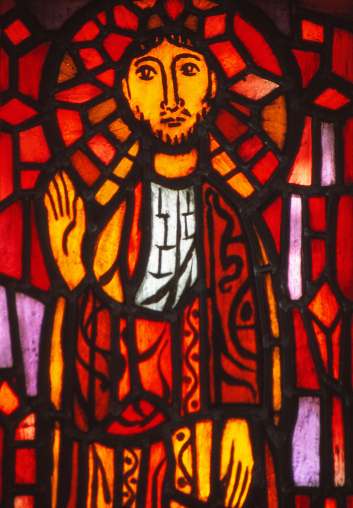 Stained-glass depiction of a haloed figure in warm red and orange hues, raising one hand in blessing, surrounded by intricate geometric shapes.