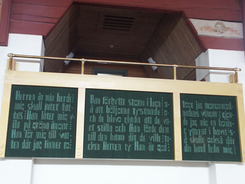 Green panels with Swedish Bible verses mounted on a gold railing inside a church balcony beneath a wooden ceiling and architectural beams.