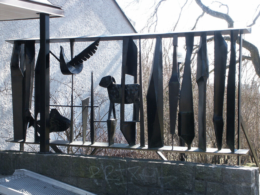 Outdoor black metal sculpture with abstract vertical shapes, a bird figure, and a stylized animal set atop a stone wall near trees.