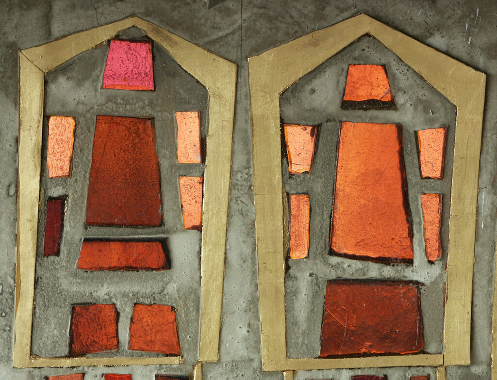 Close-up of stained-glass panels with red, orange and pink geometric pieces framed in gold on a textured concrete background.