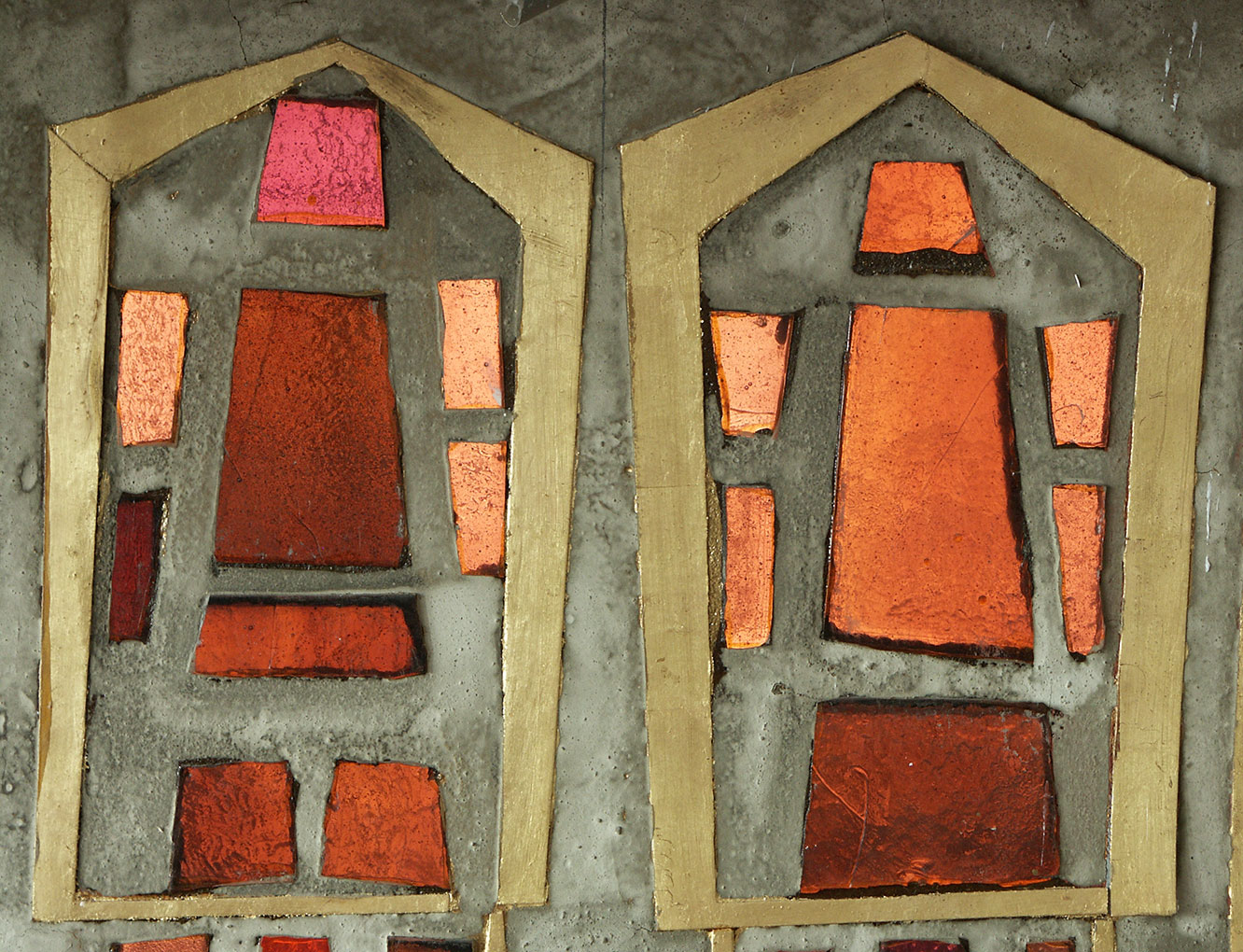 Close-up of stained-glass panels with red, orange and pink geometric pieces framed in gold on a textured concrete background.