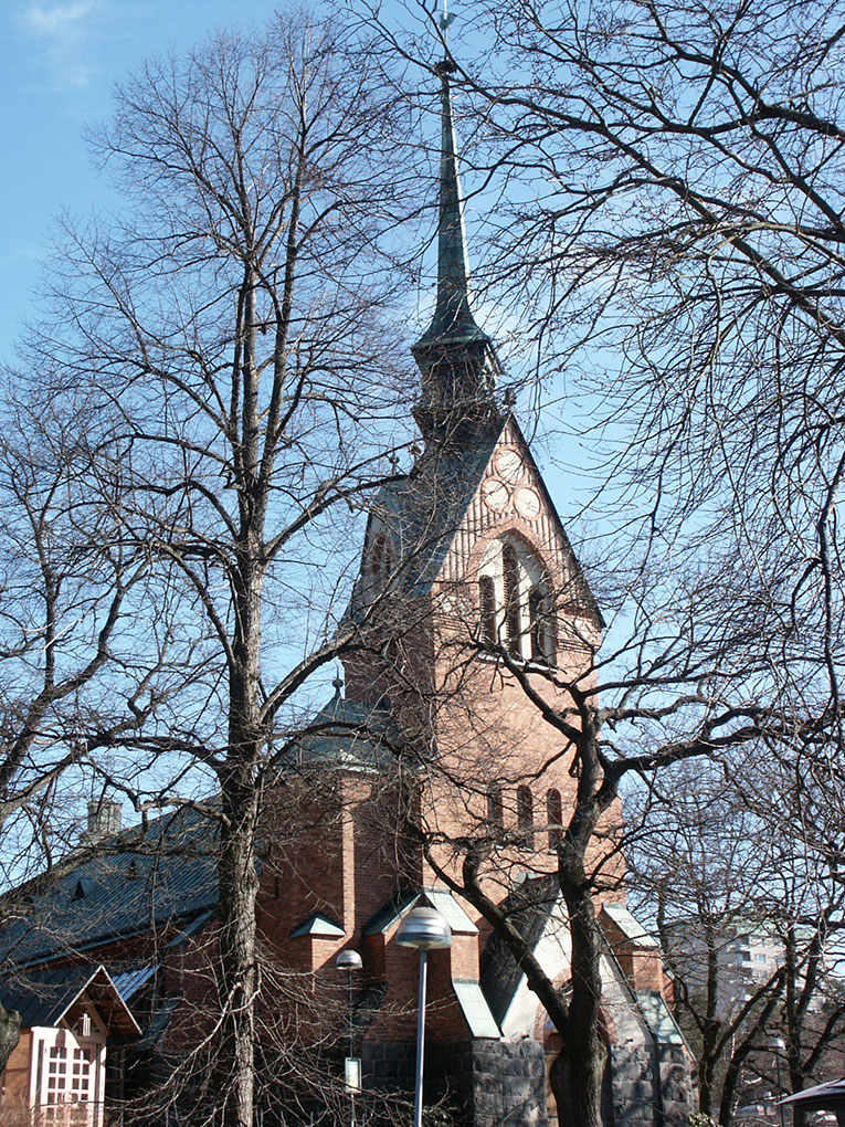 Red-brick church with a tall spire seen through leafless trees on a clear day, showing arched windows, stone base, and surrounding urban setting.
