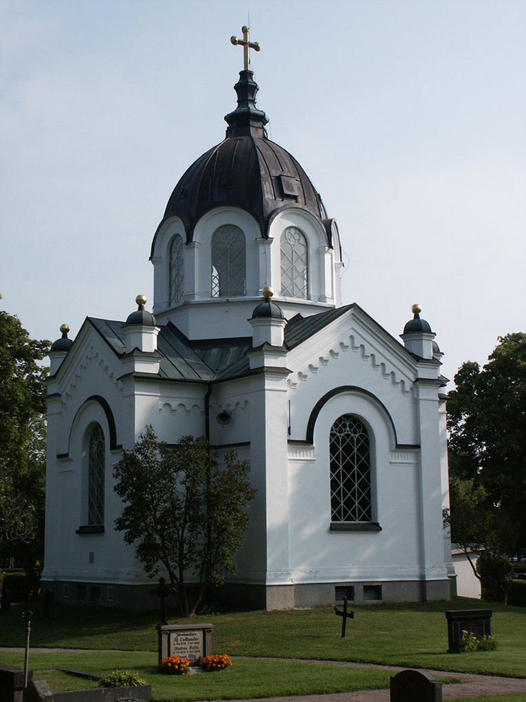 White church with arched windows, ornate trim, and a large dark dome topped by a cross, surrounded by a green churchyard and trees.
