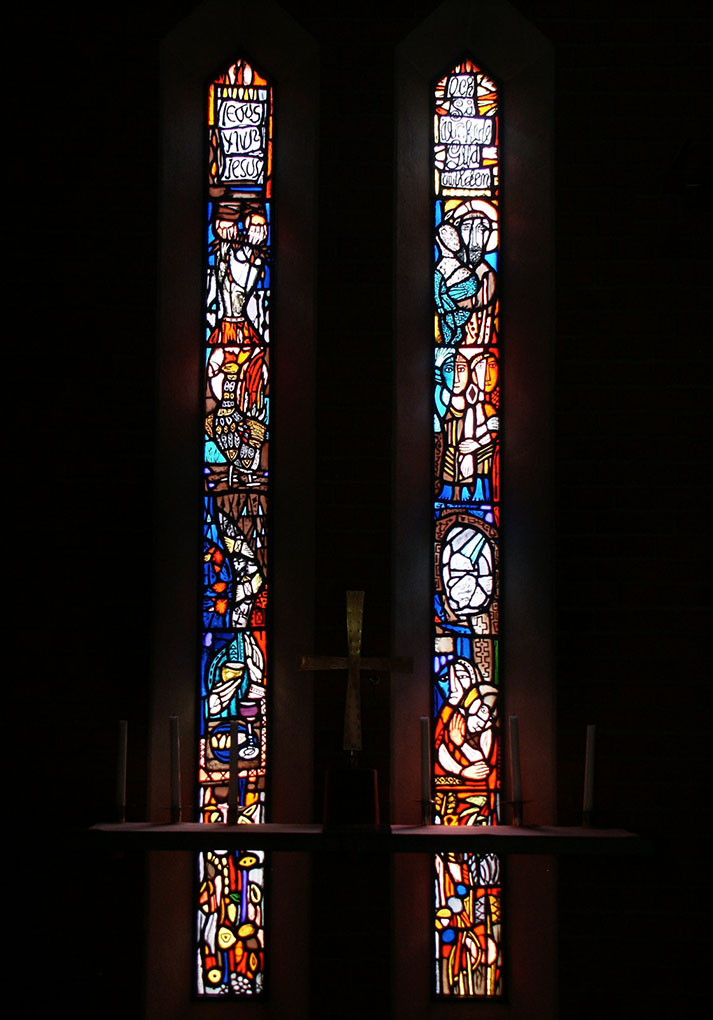 Two tall stained-glass windows filled with colorful abstract biblical scenes flank a simple altar with a metal cross and unlit white candles.
