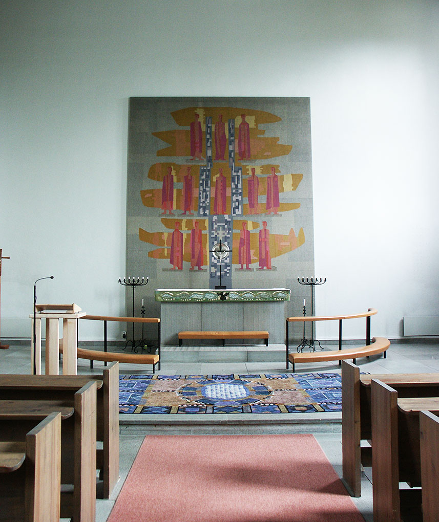 Church interior with wooden pews, altar, large abstract tapestry behind it, candlesticks, and a patterned blue carpet.