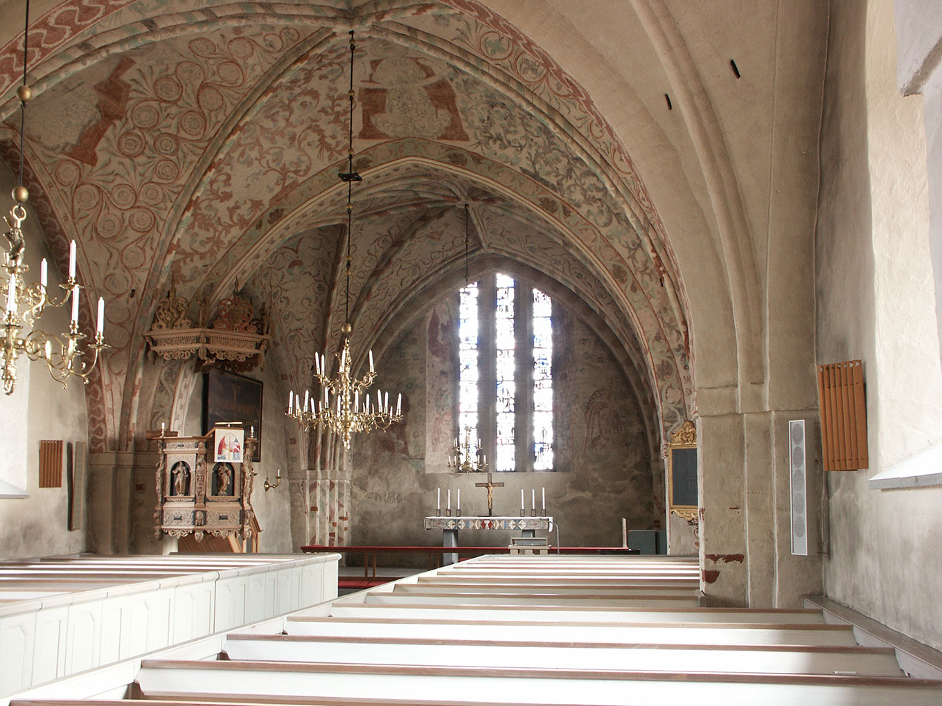004-Björksta Historic church interior with vaulted, frescoed ceilings, chandeliers, wooden pews, an ornate pulpit, and tall stained-glass windows behind the altar.