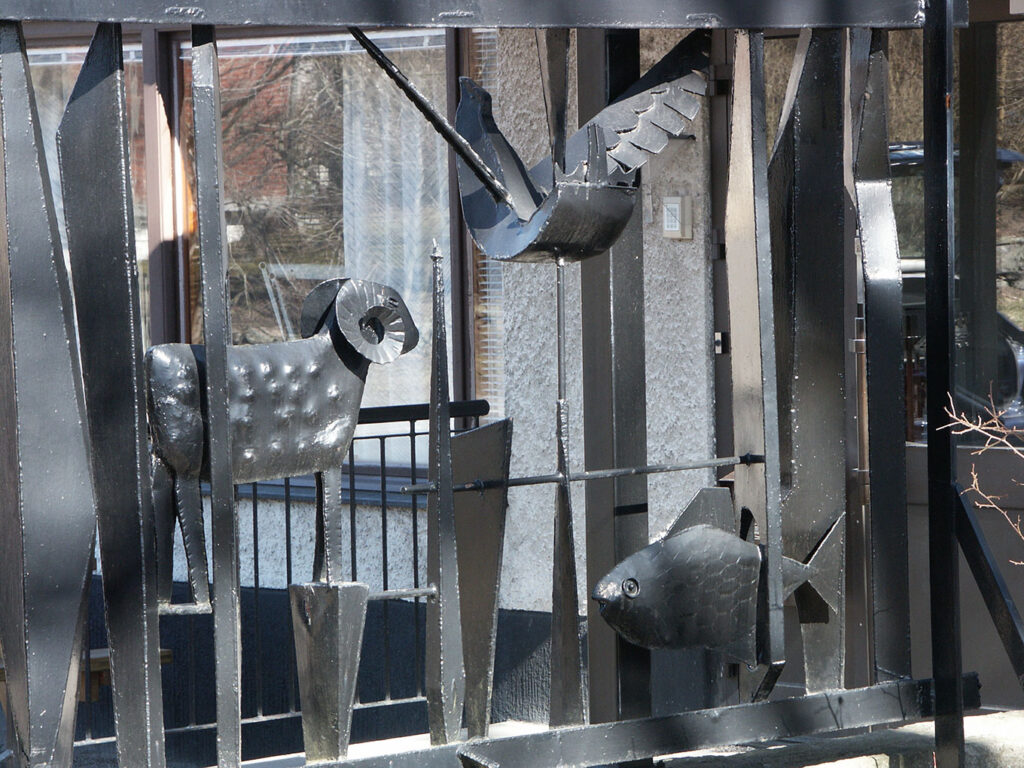 Close-up of black metal sculpture showing a stylized ram, bird in flight, and fish, framed by tall abstract vertical shapes outdoors.