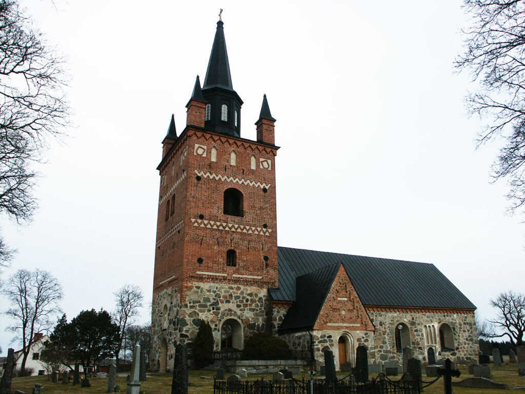 Historic brick and stone church with a tall square tower, black spire, arched windows, and surrounding graveyard under an overcast sky.