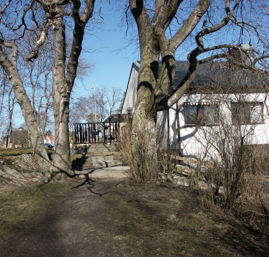 Path leading to a white building partly hidden by large leafless trees, with a black metal sculpture visible near the entrance.