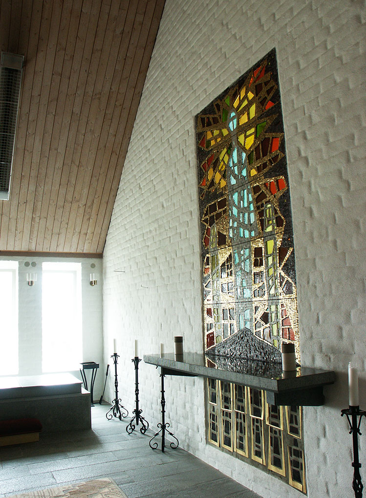 Chapel interior with tall stained-glass panel on a white brick wall, altar shelf below, and wrought-iron candle stands along the stone floor.