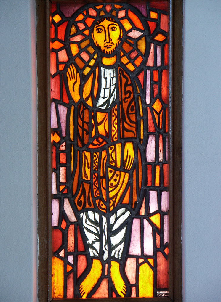 Stained-glass figure with a radiant halo, dressed in flowing robes of orange and gold, raising one hand in blessing against geometric patterns.