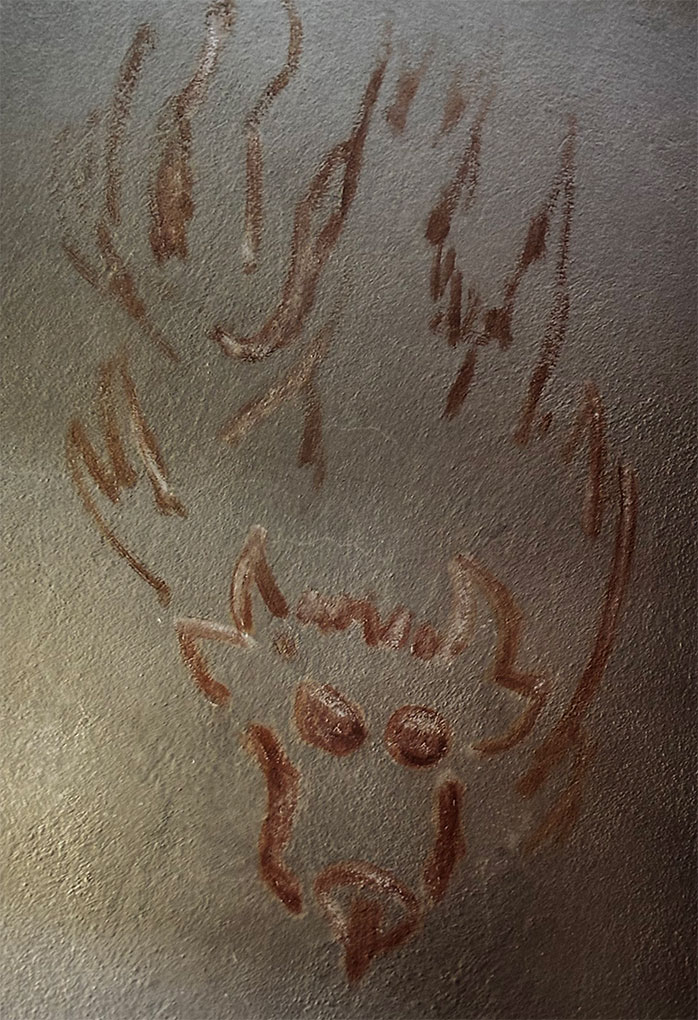Stylized brown line drawing of a horned animal face on textured gray surface, with abstract flame-like strokes rising above the figure.