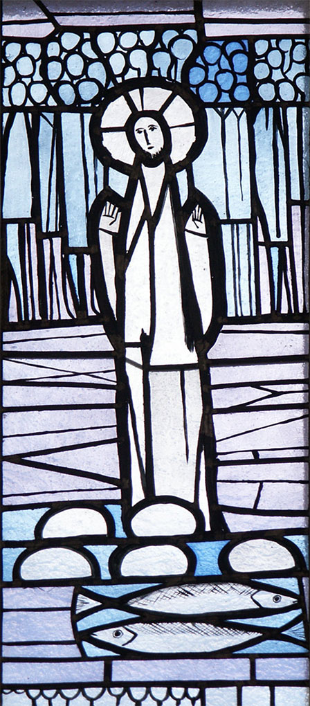 Stained-glass panel showing a haloed figure standing above loaves and fish, with stylized trees and blue geometric backgrounds.