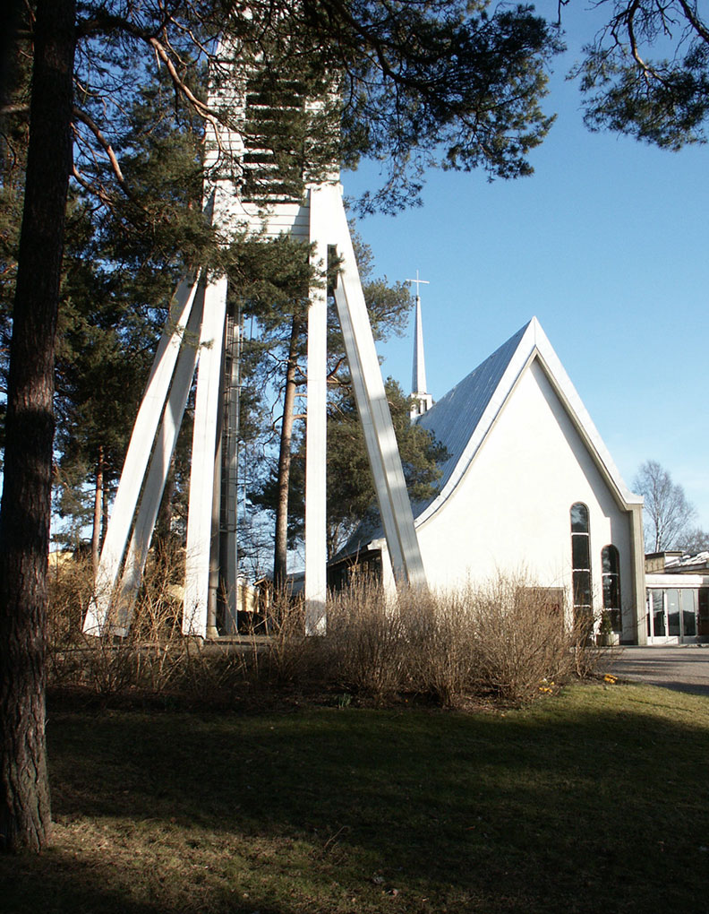 White church with tall, modern white bell tower framed by pine trees on a sunny day, with clear blue sky and surrounding bushes.
