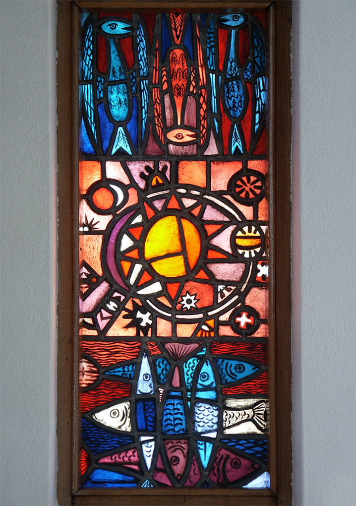 Stained-glass window featuring blue and red birds above a radiant sun motif, with multicolored fish swimming beneath in bold geometric patterns.