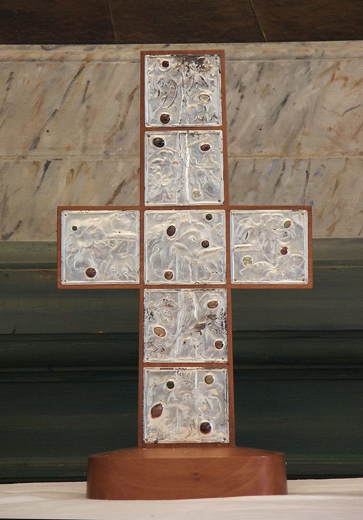 Decorative metal and wood cross composed of textured square panels with small colored stones, displayed indoors against a marble and dark green backdrop.