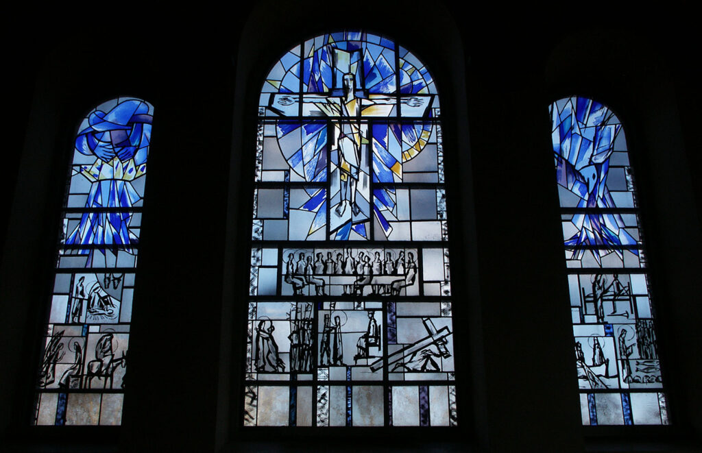 Three stained-glass windows depicting abstract blue and yellow figures above detailed biblical scenes rendered in bold black line art.