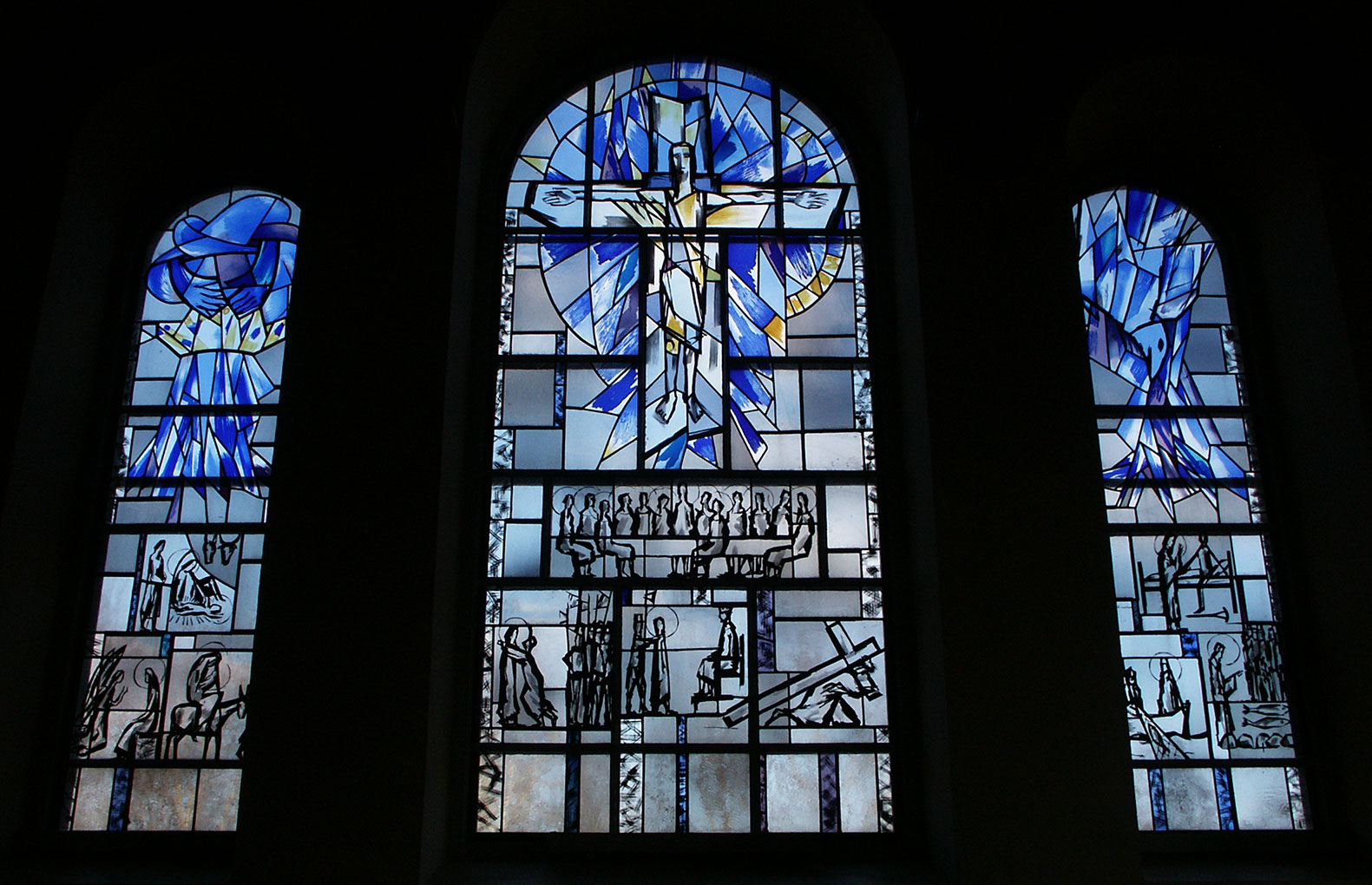 Three stained-glass windows depicting abstract blue and yellow figures above detailed biblical scenes rendered in bold black line art.