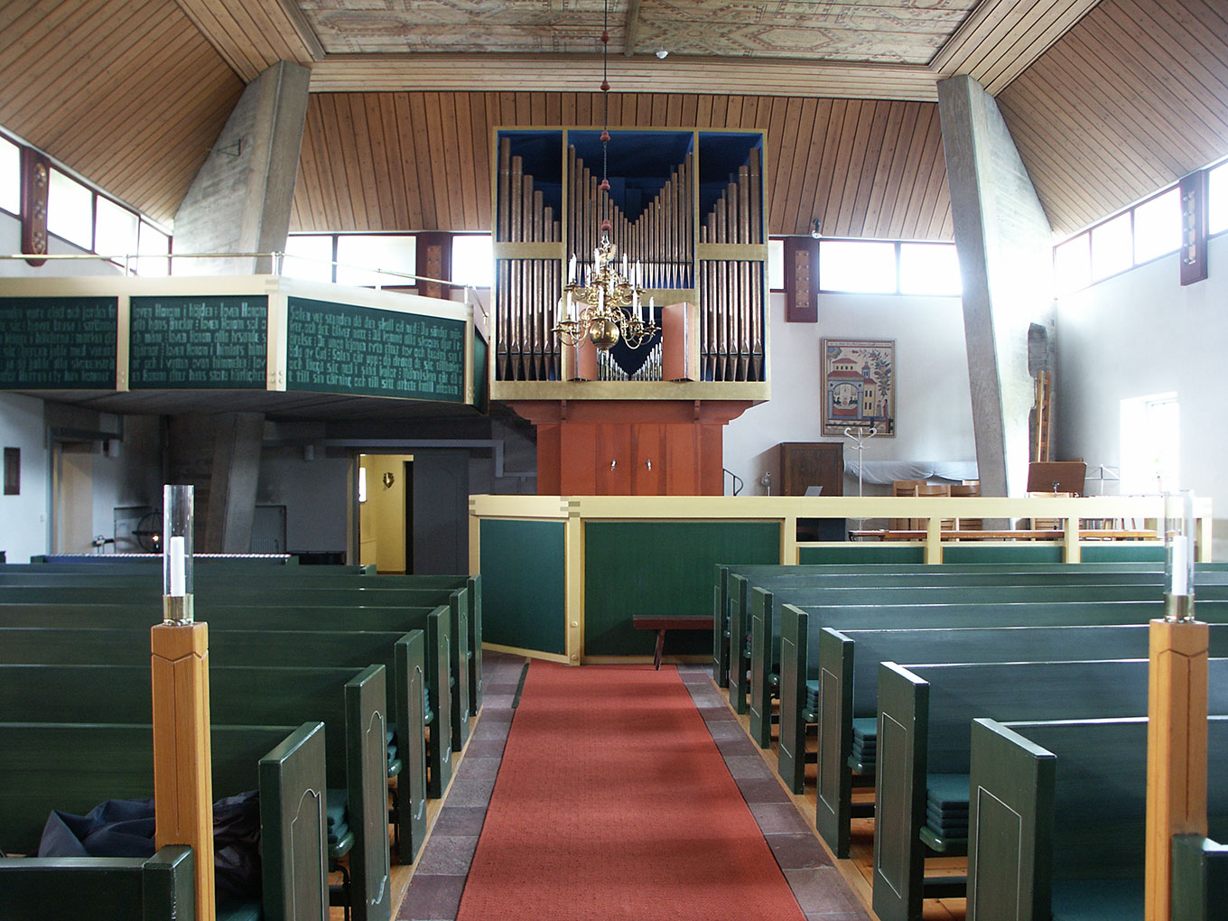 A wide view of a modern church interior with green pews, a red carpeted aisle, and a large pipe organ on the rear balcony under a wood-paneled ceiling.