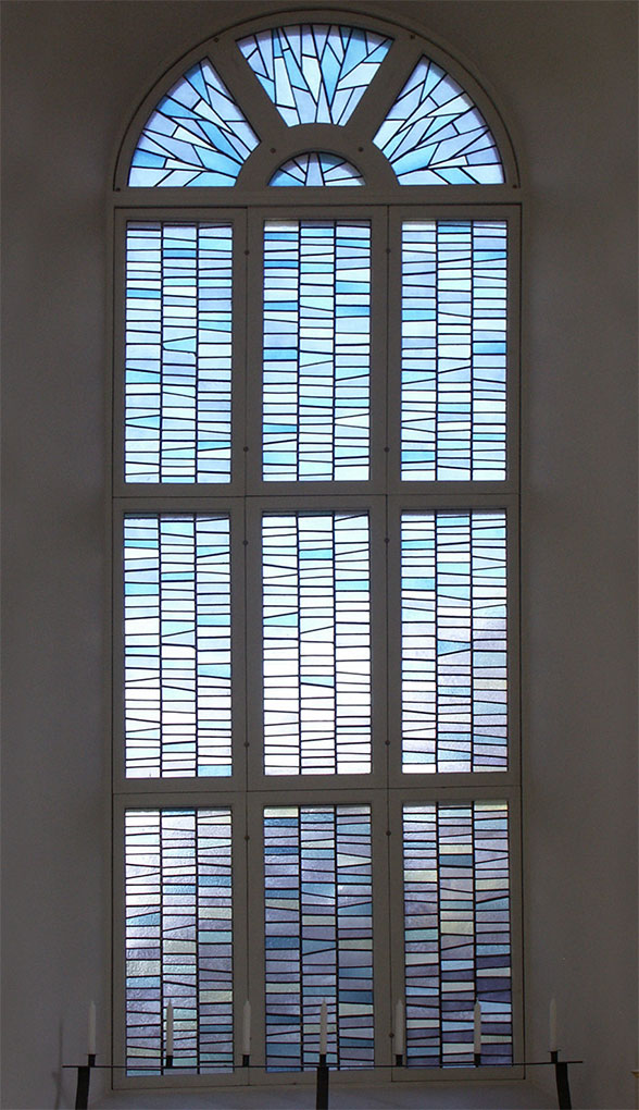 Tall arched stained-glass window with repeating blue geometric panes arranged in vertical sections, creating a soft, luminous patterned glow.