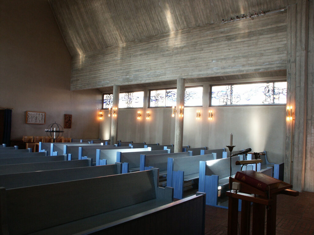 Blue-pewed church interior with high wooden ceiling, narrow stained-glass clerestory windows, wall sconces, and a lectern in the foreground.