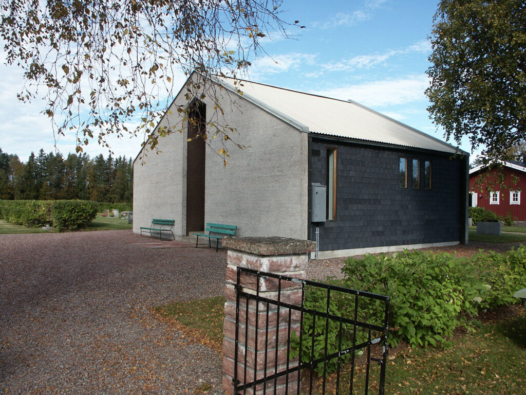 Small modern chapel with grey walls and a white roof, set in a peaceful cemetery with benches, trees, shrubs, and a brick-and-iron gate.