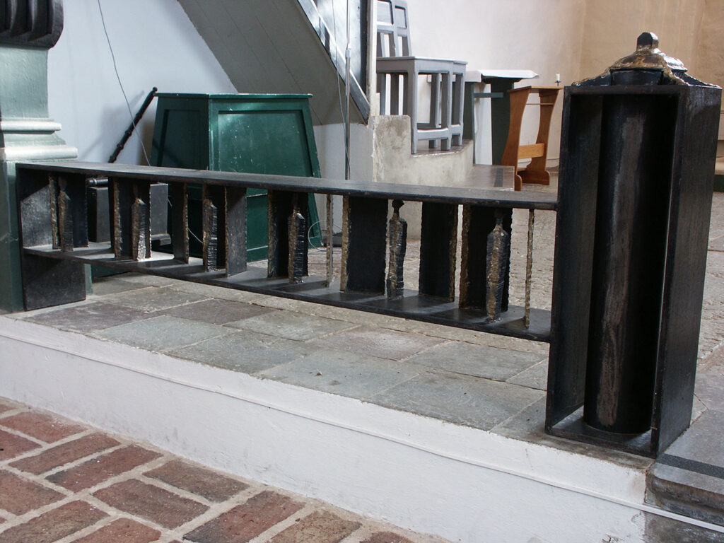 Dark metal altar rail with vertical sculpted elements on a stone step inside a church, surrounded by wooden furniture and light-toned walls.