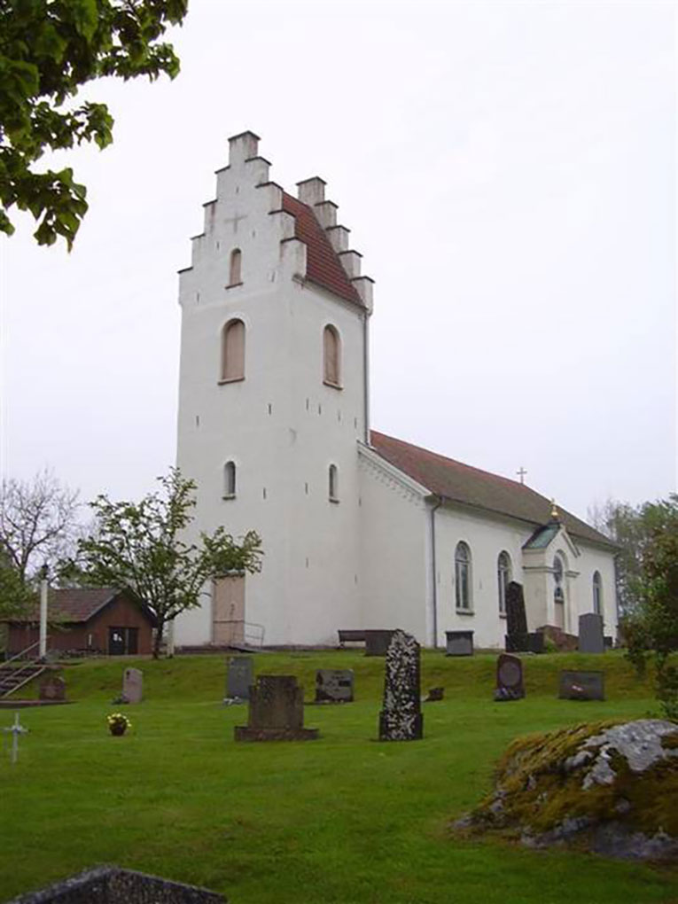 White church with a stepped tower and red-tiled roof, surrounded by a green churchyard dotted with gravestones and trees.