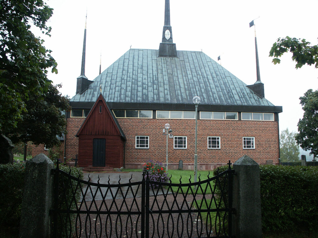 Red-brick church with tall central spire and smaller corner spires, fronted by a wooden porch and black gate, trees framing the scene.