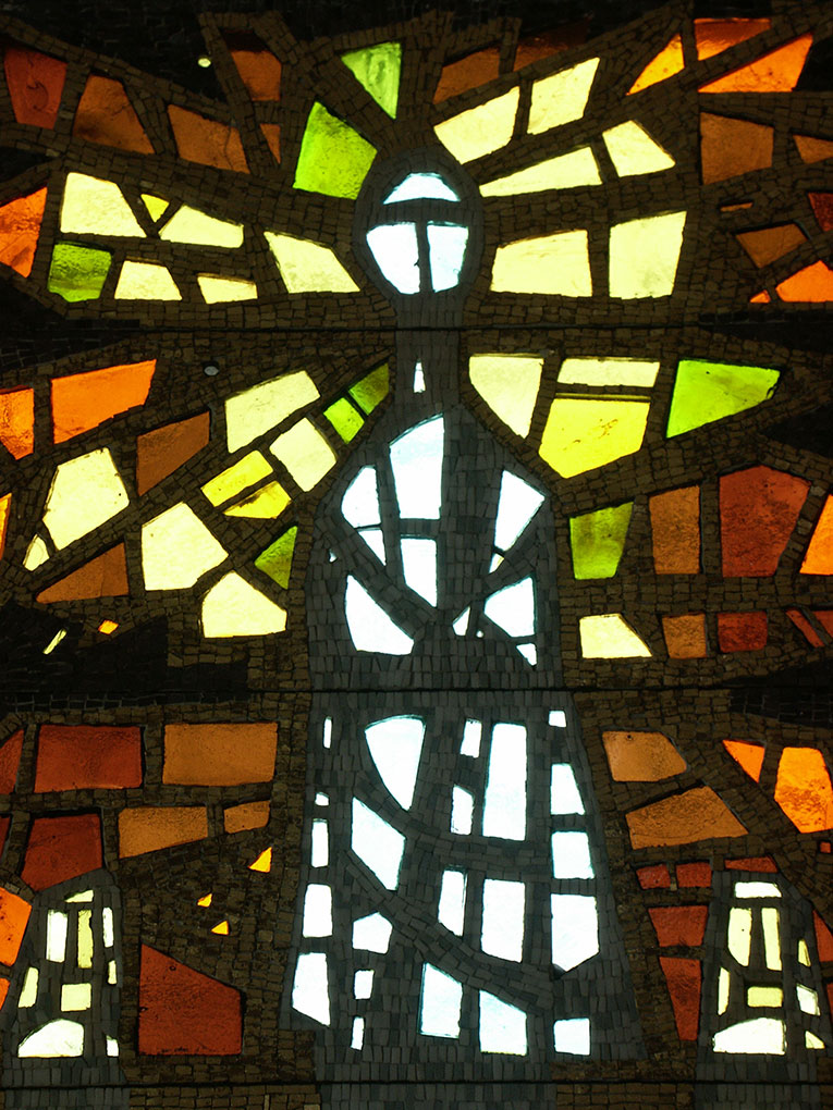 Stained-glass mosaic featuring a tall, glowing central figure in cool tones surrounded by radiant yellow, green, and orange geometric shards.
