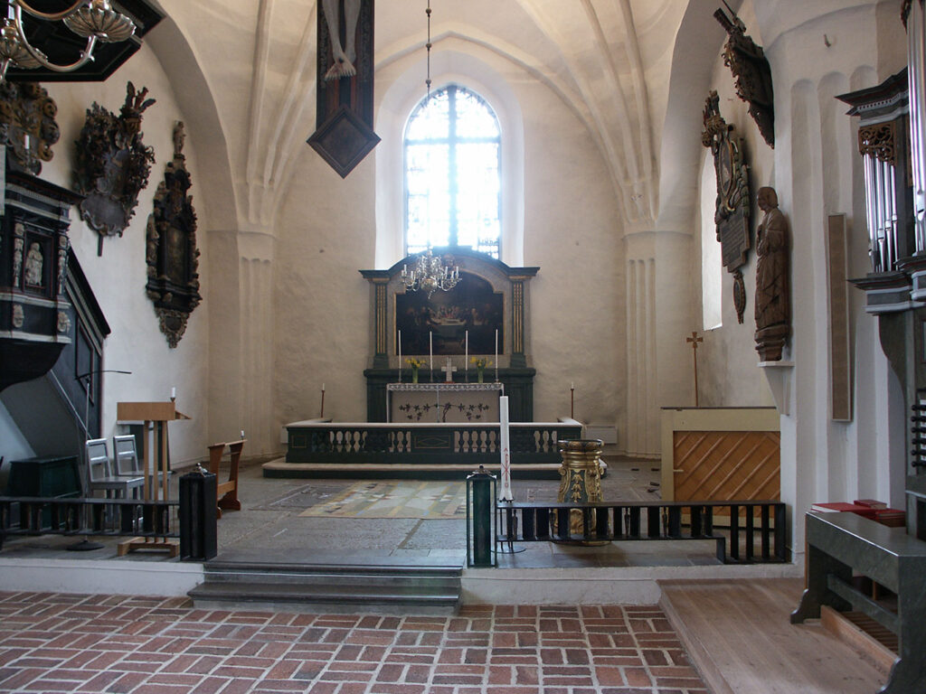 Church altar with green cloth, framed painting, brass chandelier, and tall stained-glass window illuminating the arched sanctuary interior.