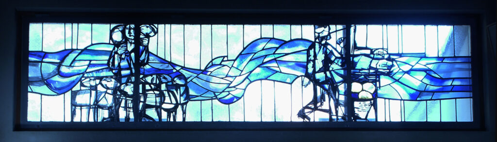 Vibrant blue stained-glass window with abstract figures, flowing waves, and symbolic shapes arranged across a wide horizontal panel.