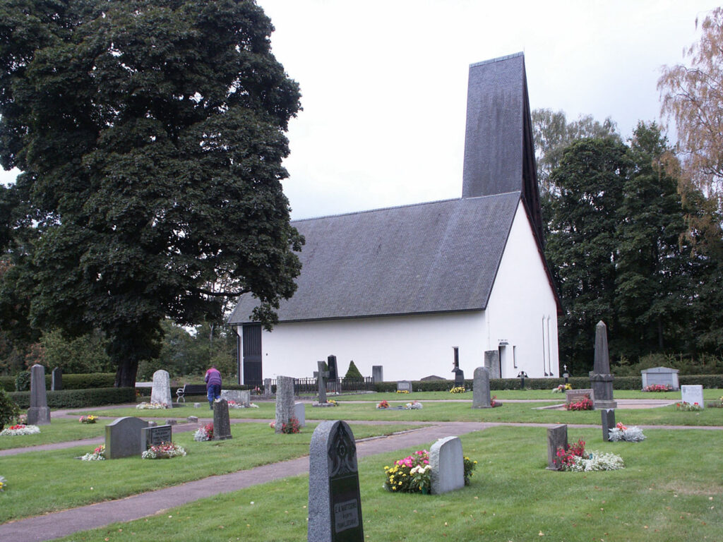 White church with a steep dark roof and tall tower rising above a peaceful graveyard filled with headstones, trees, and neatly kept grass.