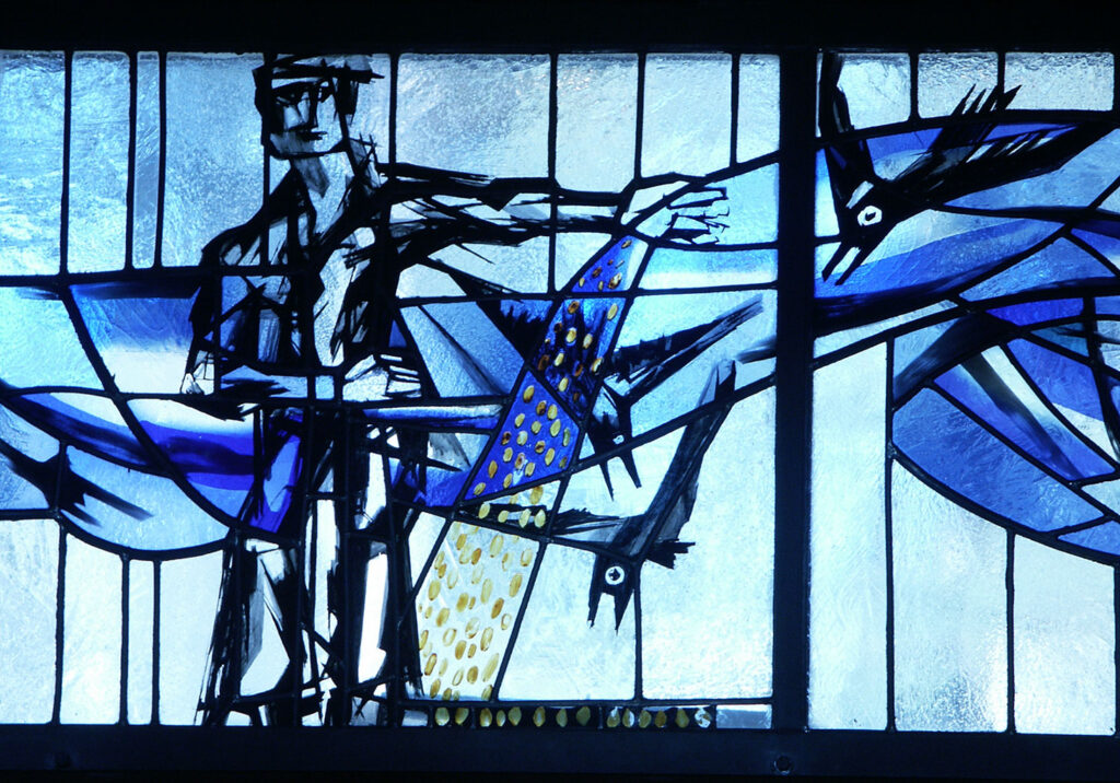 Blue-toned stained glass panel showing a stylized figure scattering seed while black birds swoop nearby against abstract flowing shapes.