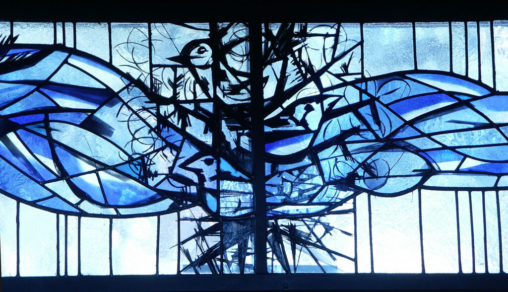 Stained glass window with abstract blue shapes and black outlines forming a tree-like composition with birds and flowing wave-like patterns.