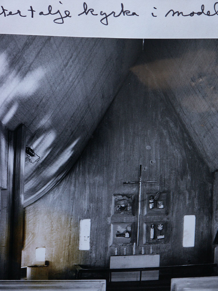Black-and-white photo of a church interior with wooden walls, a simple cross, small wall reliefs, and handwritten text visible at the top.